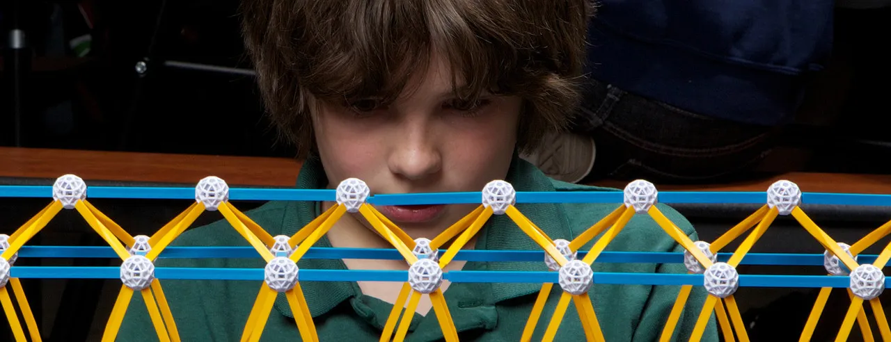 A boy looking through a long bridge he built from Zometool struts and balls
